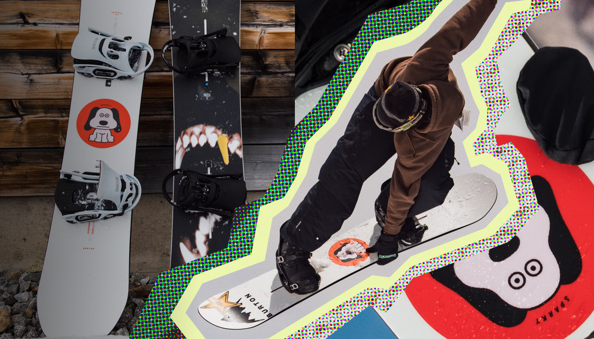 A lifestyle shot of a snowboarder in a brown jacket and black pants riding a white snowboard, with two snowboards standing vertically on a wooden deck in the background. The snowboard and boards feature a bold graphic design with a red circular logo containing a white dog head and a black-and-white shark mouth graphic.