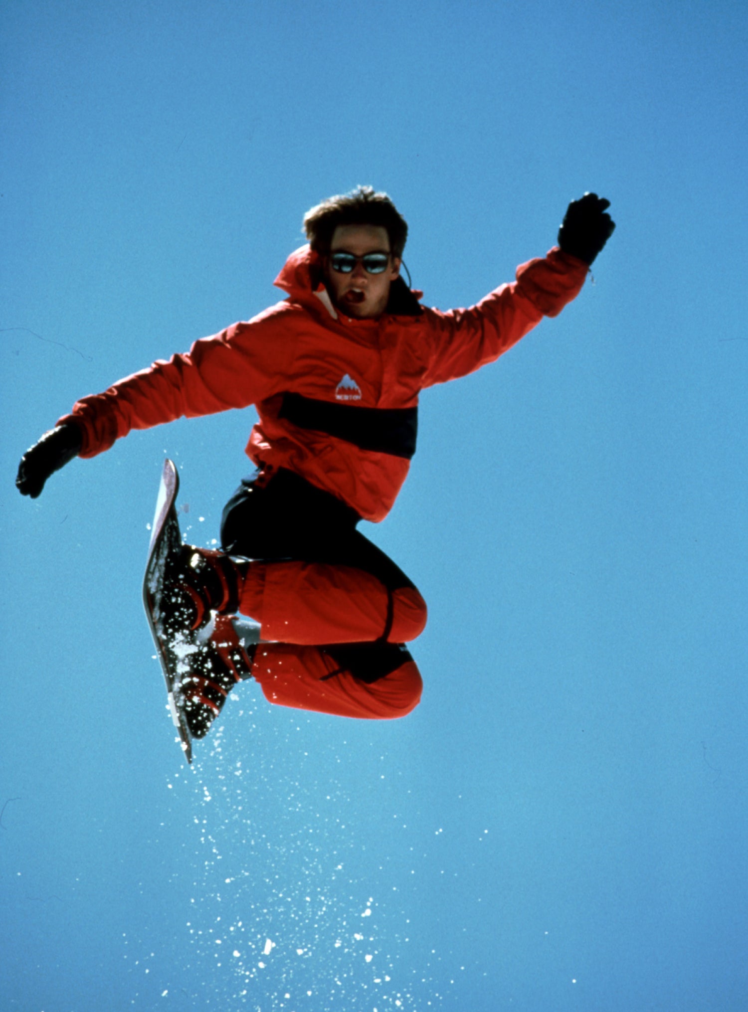 A snowboarder in a bright red and black ski suit jumps high against a clear blue sky, with snow spraying from the board.