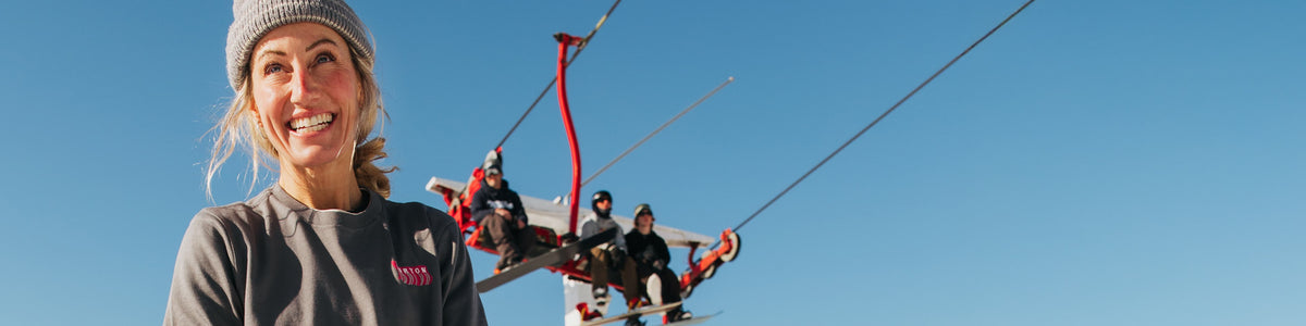 A smiling woman in a gray beanie and gray sweatshirt stands in front of a ski lift with riders on a clear blue sky background.