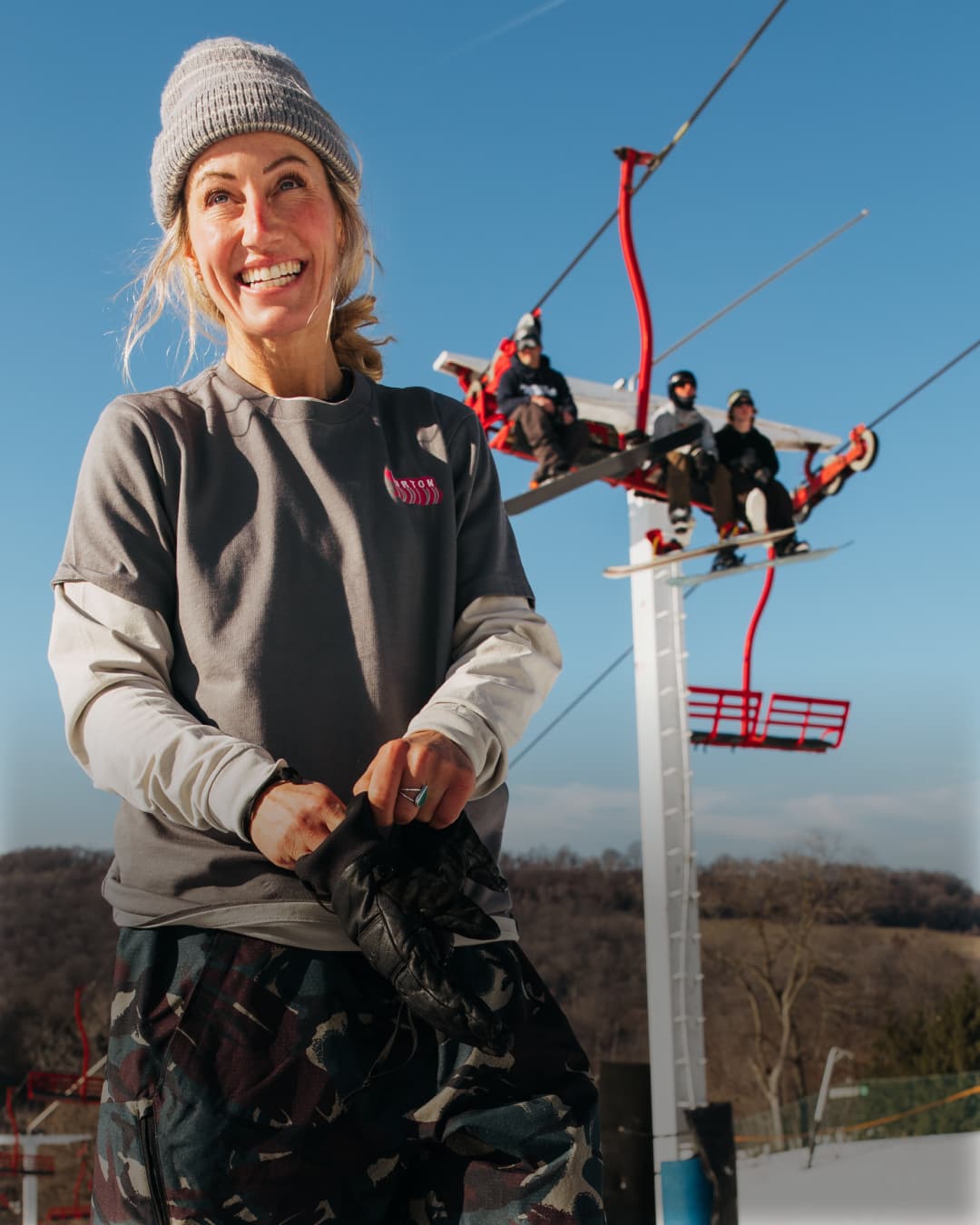 A smiling woman in a gray beanie and gray sweatshirt stands in front of a ski lift with riders on a clear blue sky background.