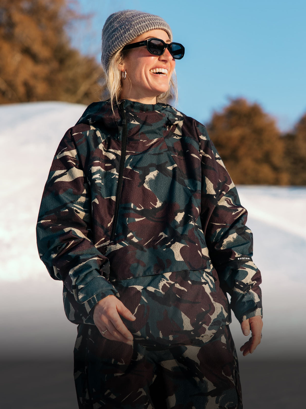 A woman wearing a camouflage hooded jacket and matching pants, a gray knit beanie, and black sunglasses, standing outdoors in a snowy setting with trees in the background.