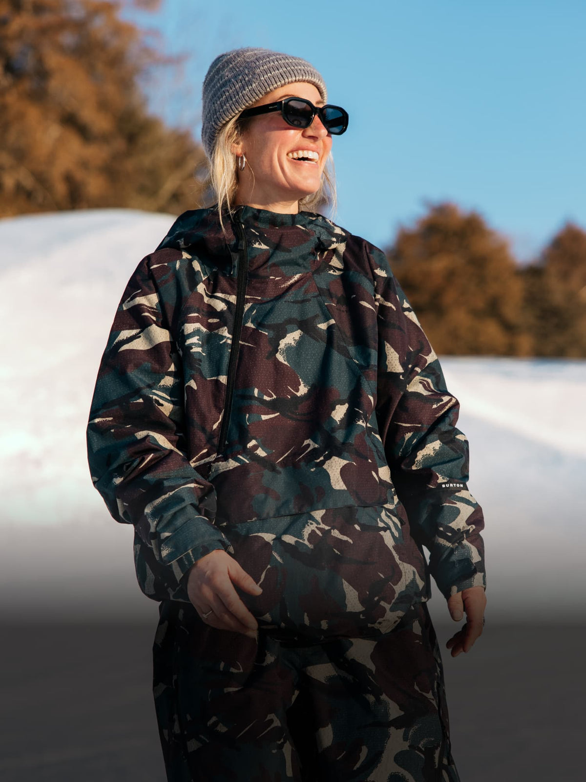A woman wearing a camouflage hooded jacket and matching pants, a knit beanie, and sunglasses, standing outdoors in front of a snowy slope and trees under a blue sky.