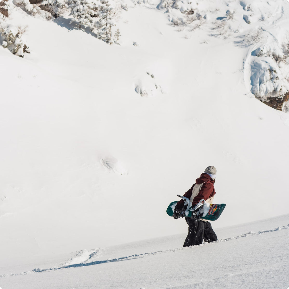 A snowboarder in a red jacket and dark pants walks through deep snow while carrying a teal snowboard, with a snowy mountain slope and rocky outcrops in the background.