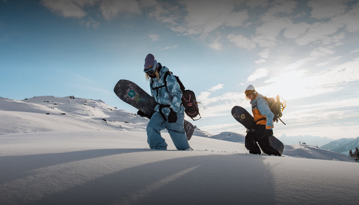 Two snowboarders in full snowboard gear walking through deep snow on a mountain slope under a bright sky with the sun breaking through clouds.
