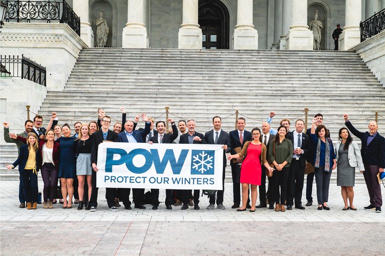 Group of people holding a 'POW Protect Our Winters' banner on steps of a government building.
