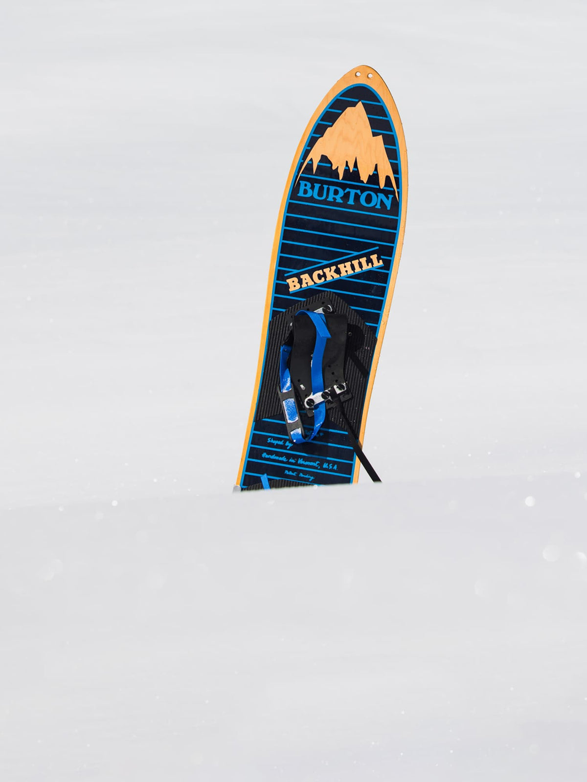 A Burton Backhill backboard standing upright in snow, featuring a blue and black striped design with an orange mountain graphic and Burton branding.