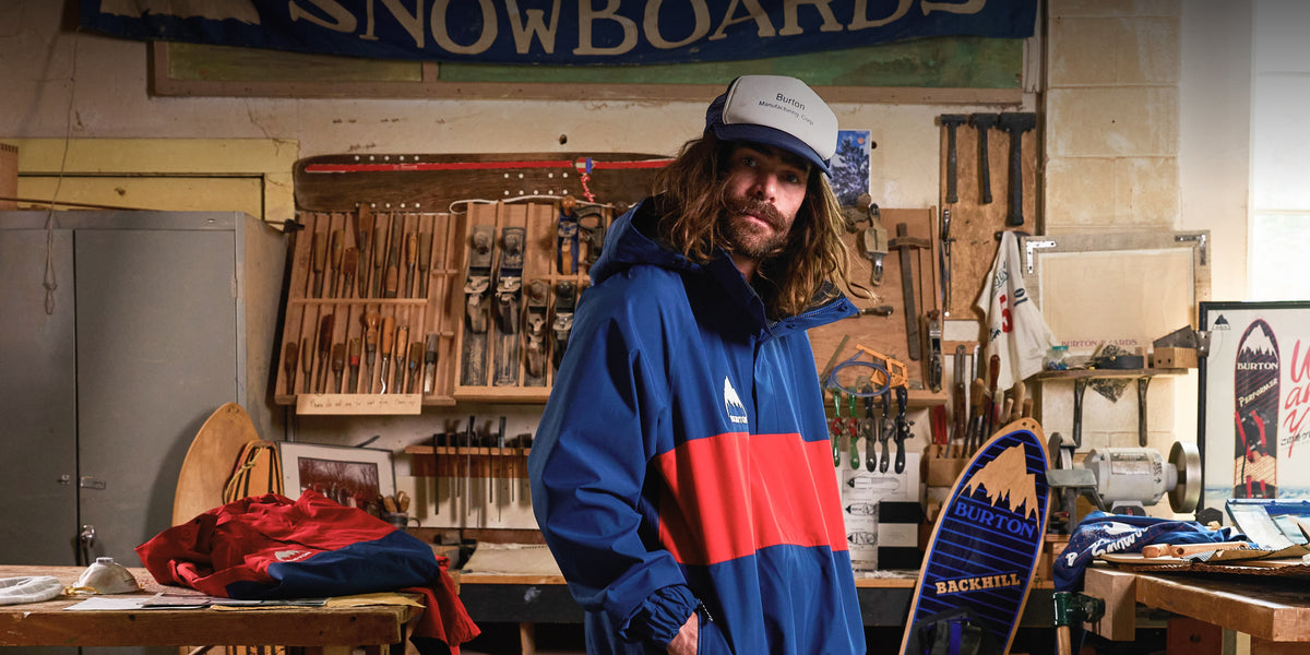 A man with long hair and a beard wearing a blue, red, and white color-blocked snowboard jacket and a white cap, standing in a workshop filled with tools and equipment.