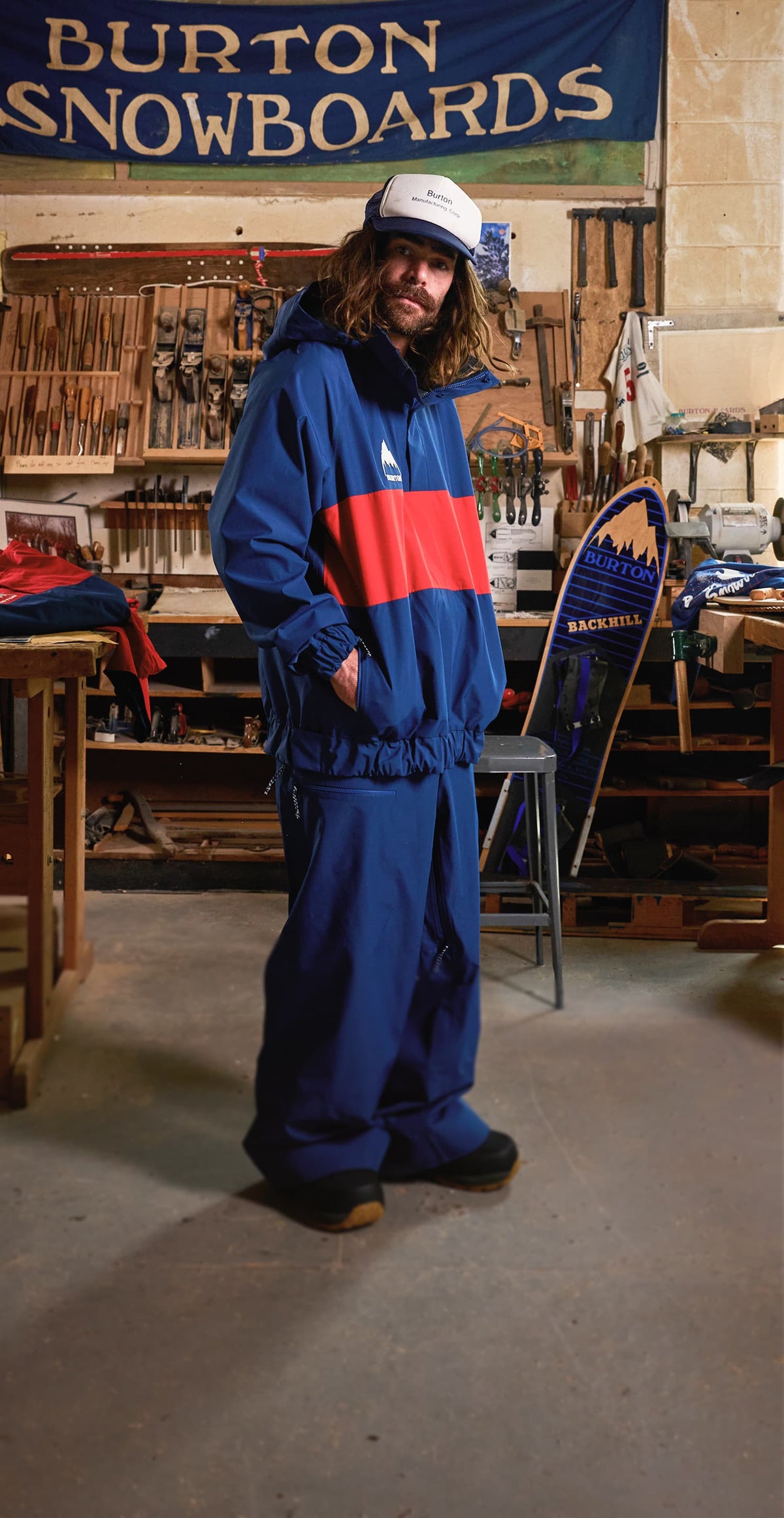 A man wearing a blue, red, and white color-blocked Burton snowboard outerwear stands in a workshop filled with snowboards and apparel. The outerwear features a blue base with a wide red band and a white Burton logo on the chest.