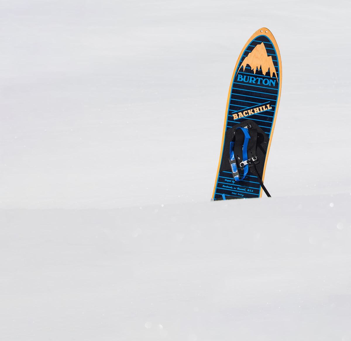 A Burton Backhill snowboard standing upright in deep snow, showing its blue, black, and orange graphic design.
