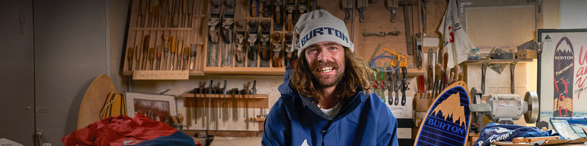 A smiling man wearing a white beanie with 'BURTON' printed on it and a blue and red jacket stands in a workshop filled with vintage skis and wooden tools. A blue and yellow 'BACKHILL' board is held up on the right.
