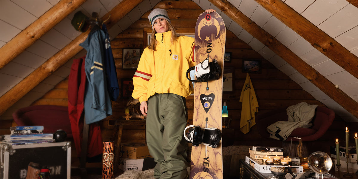 A woman wearing a yellow jacket, olive green snow pants, and a gray beanie stands indoors holding a Burton snowboard. The snowboard has a wood-grain pattern with black bindings and visible Burton branding.