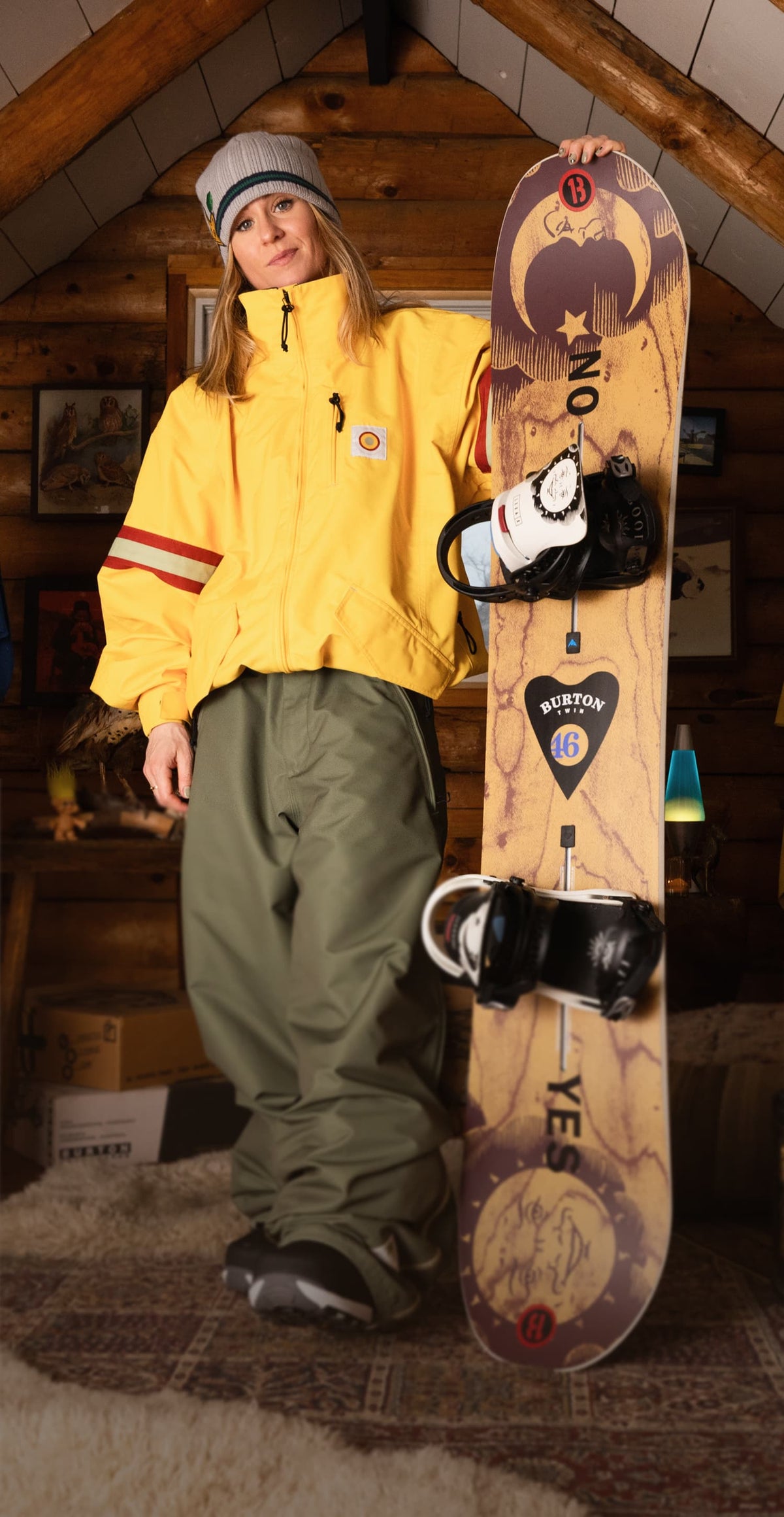 A woman wearing a yellow jacket, olive green snow pants, and a gray beanie stands indoors holding a Burton snowboard. The snowboard has a wood-grain pattern with black bindings and visible Burton branding.