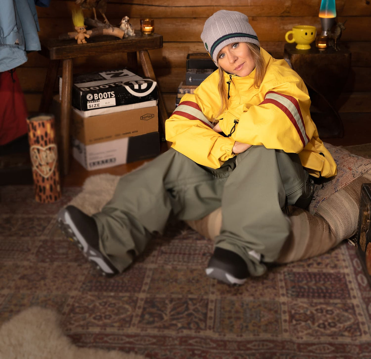 A person sitting on a patterned rug in a rustic cabin interior, wearing a bright yellow snowboard jacket with red and white stripes, olive green snow pants, and a gray beanie.