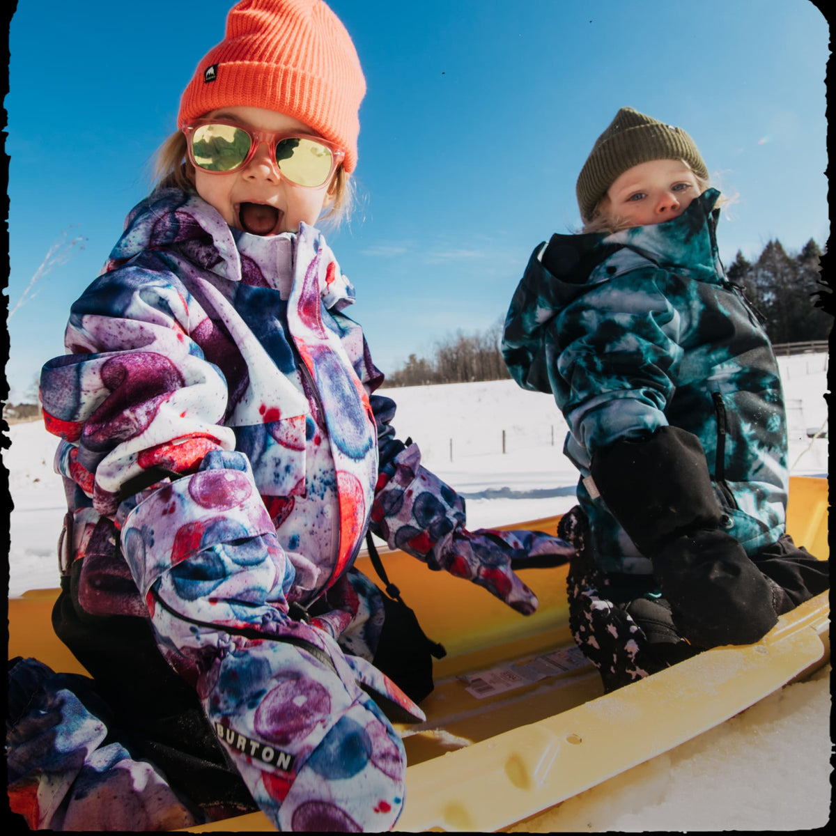Two children wearing colorful winter jackets and hats sitting in a yellow sled outdoors on a snowy day under a blue sky. The child in front wears an orange beanie, gold-tinted sunglasses, and a multicolor patterned snow jacket with a Burton logo near the bottom left hem.