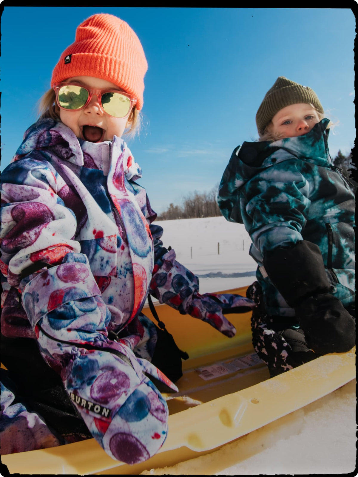 Two children wearing winter gear, including colorful snow jackets, beanies, and sunglasses, sitting in a yellow sled outdoors in the snow under a clear blue sky.