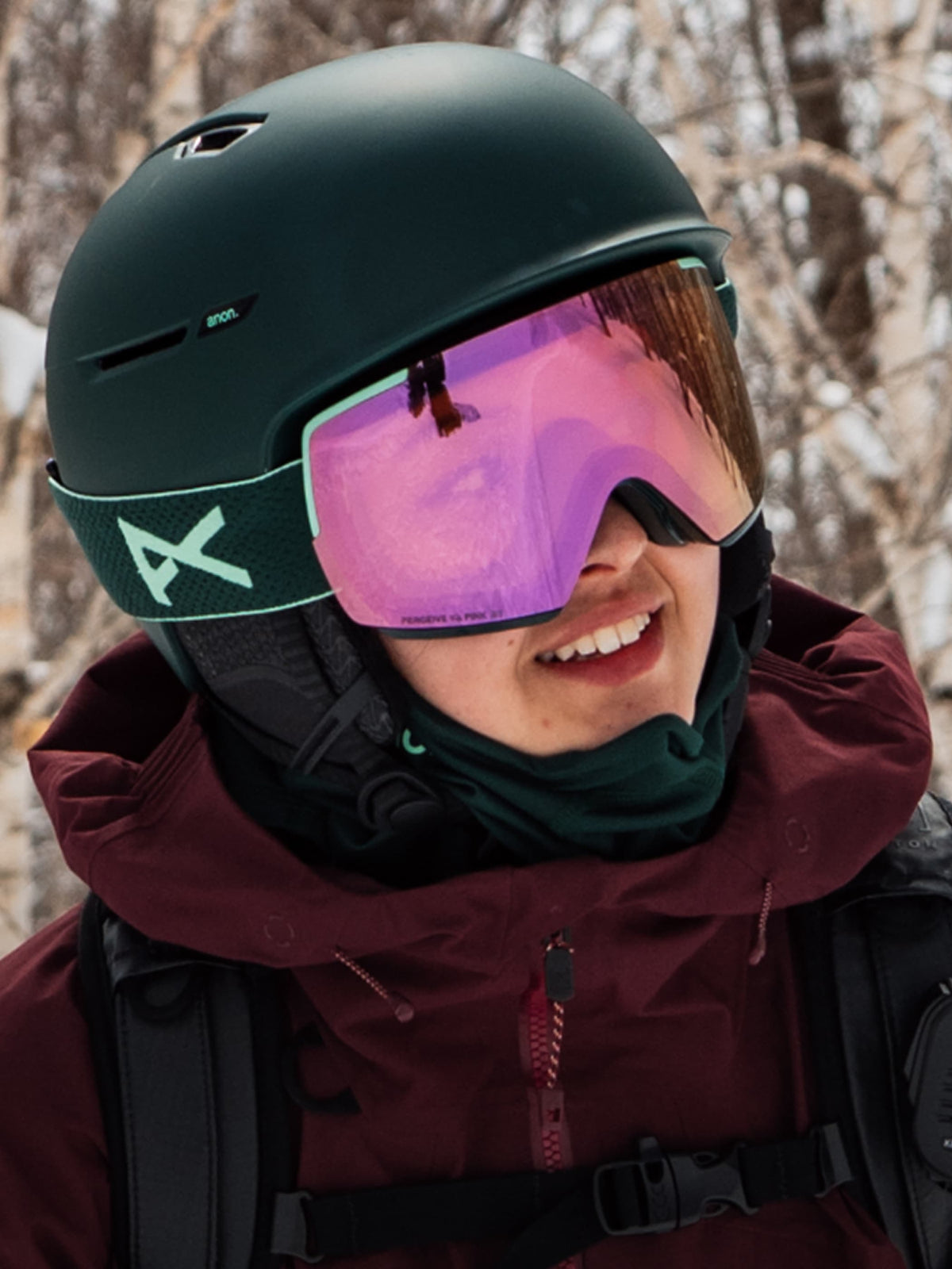 A close-up of a person outdoors in winter gear, wearing a matte black ski helmet with a green-accented strap, pink-tinted ski or snowboard goggles, a dark green neck gaiter, and a burgundy jacket.