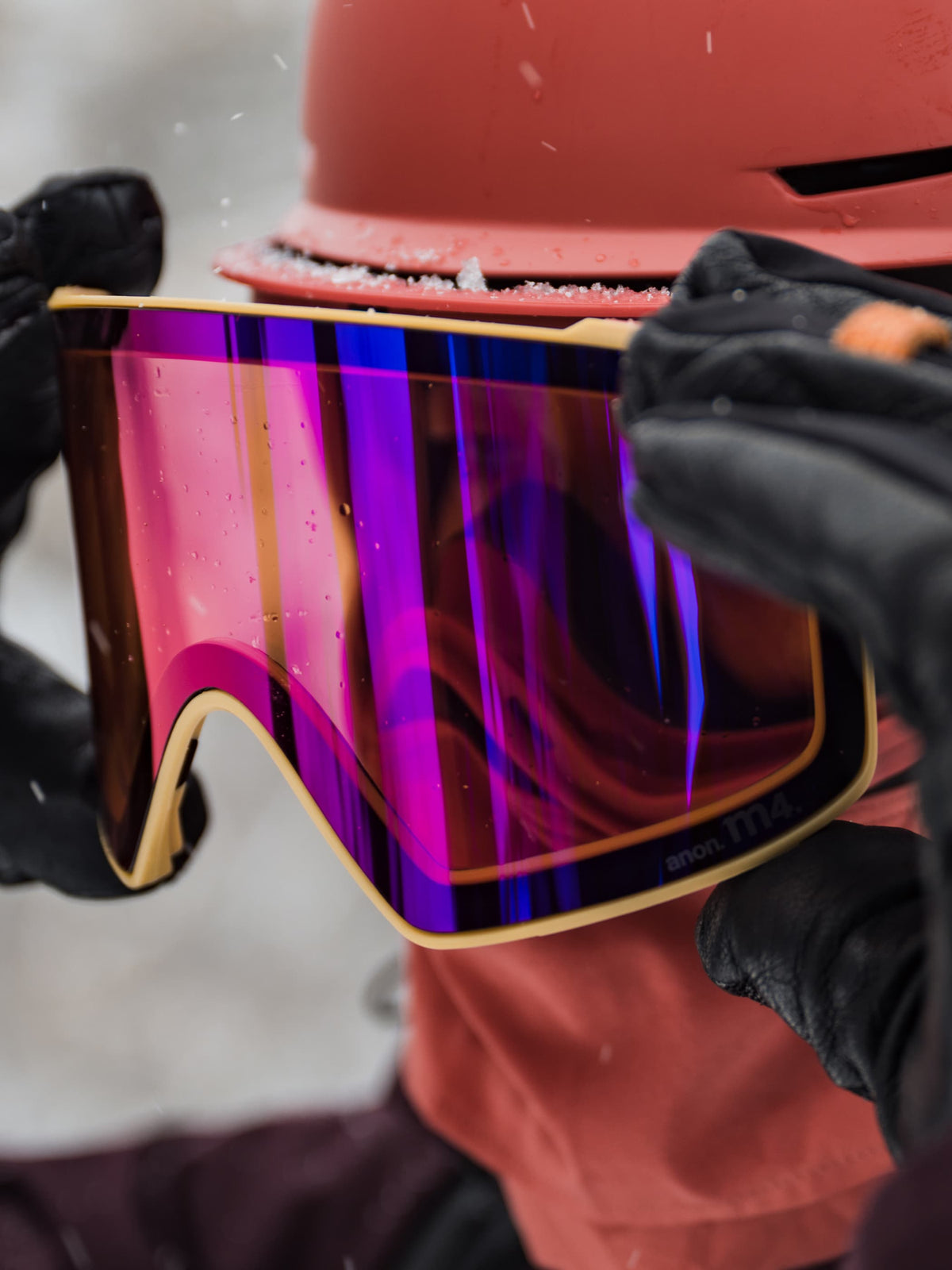 Close-up of a person wearing black gloves and a coral-orange helmet while holding a ski or snowboard goggles with a large mirrored purple, pink, and blue lens and a gold frame.