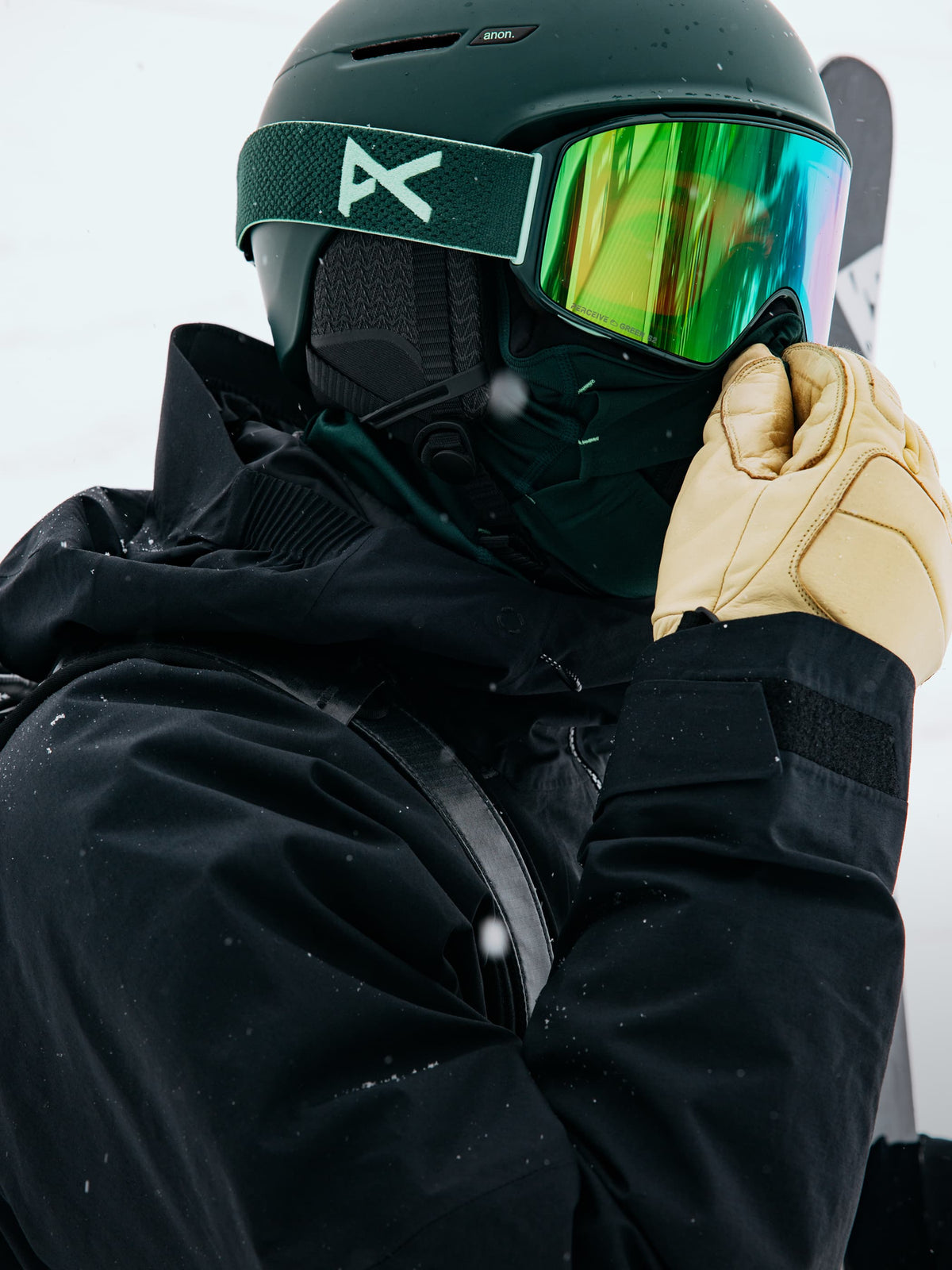 A close-up of a snowboarder wearing a dark green helmet with a reflective green and gold mirror lens, a black ski jacket, and a beige glove, set against a snowy white background.