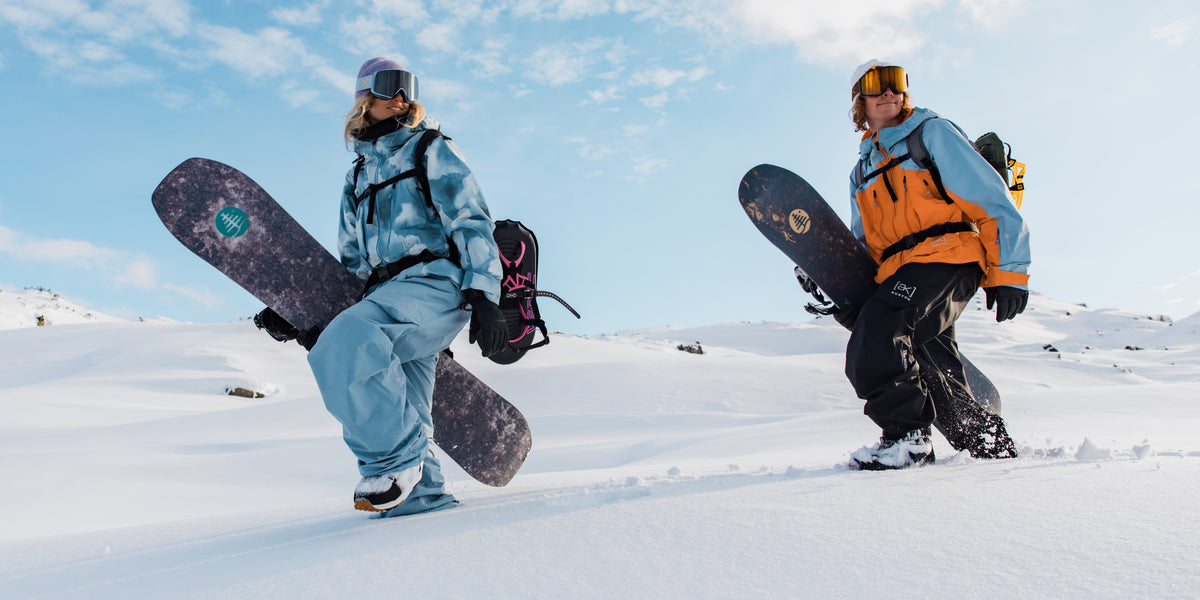 Two snowboarders posing on a snowy mountain under a blue sky. The person on the left wears a light blue snowsuit and holds a dark snowboard.