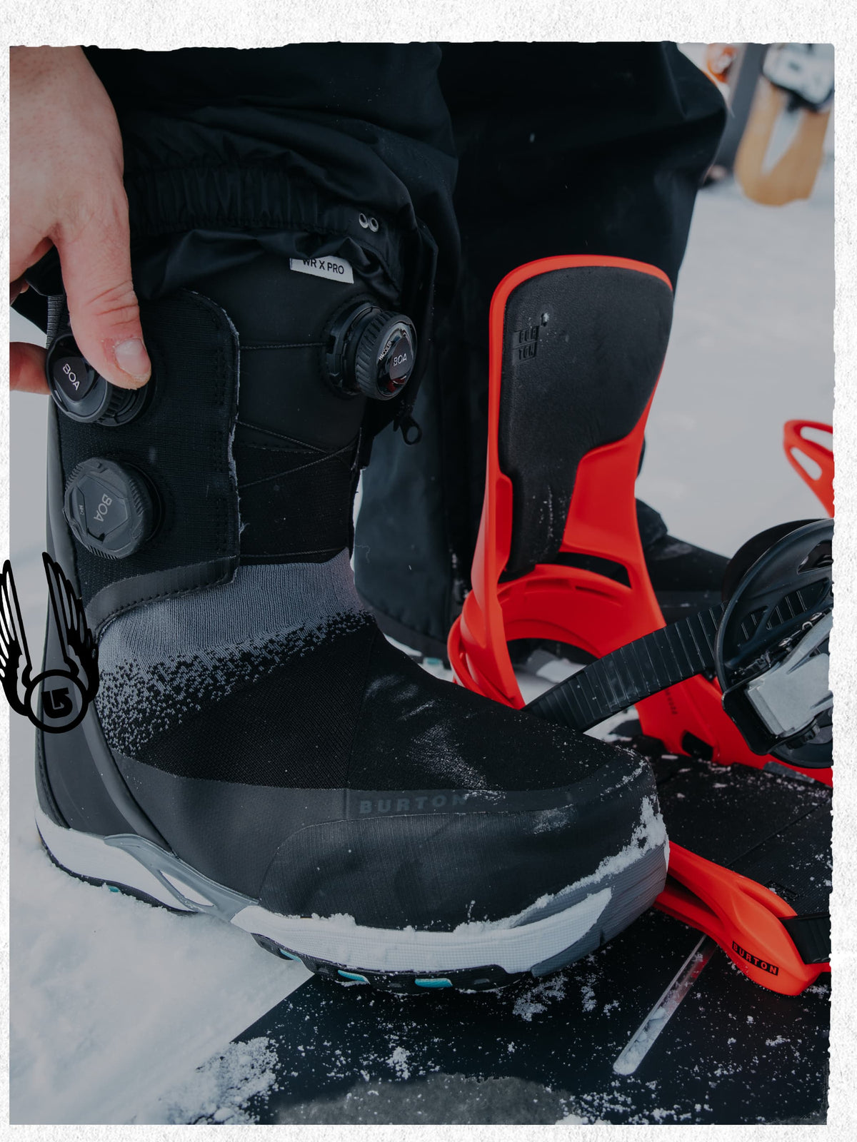 A close-up of a Burton snowboard boot with a BOA dial closure system, being adjusted by a hand on a snowboard with bright orange bindings in a snowy outdoor setting.