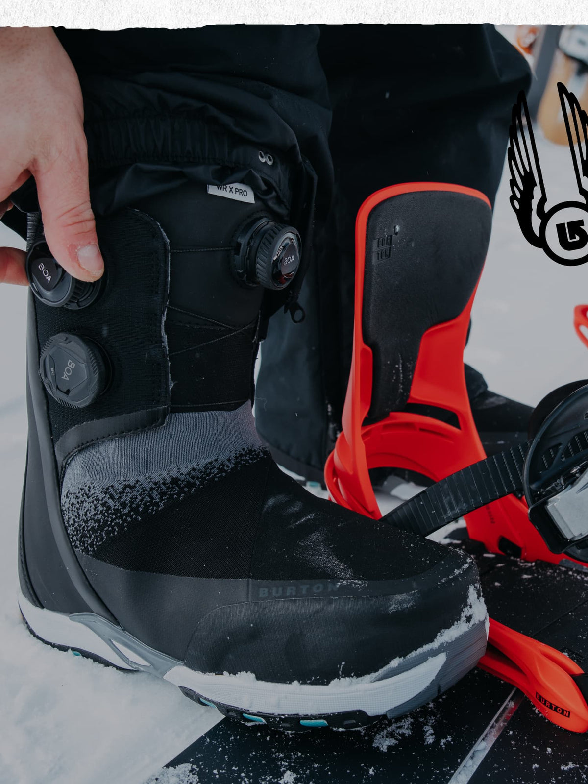 A close-up of a Burton snowboard boot with a BOA dial closure system, being adjusted by a hand on a snowboard with bright orange bindings in a snowy outdoor setting.
