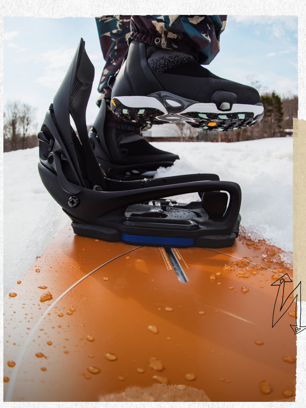 Black snowboard bindings with a blue accent, shown on a snowboard with a person's legs and boots visible in a snowy outdoor setting.