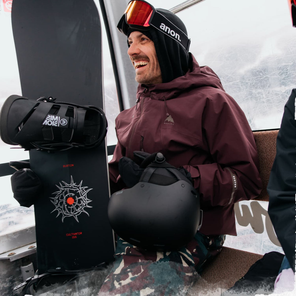 A smiling man in ski resort attire sits on a lift, holding a black snowboard with a white graphic and a black helmet. He wears a burgundy hooded jacket, black gloves, and a black beanie with orange-tinted goggles.