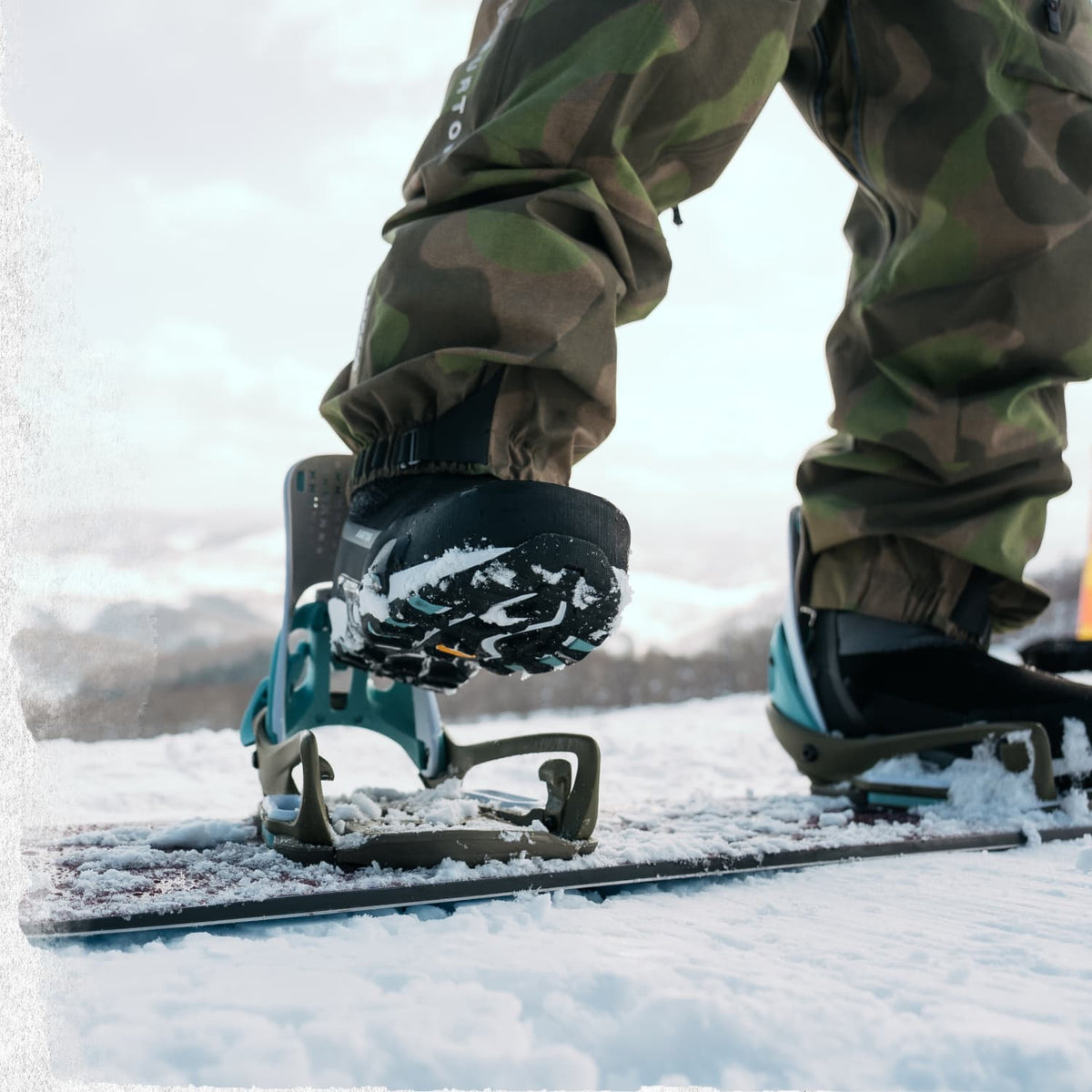 Close-up of a person snowboarding in the snow, showing the bindings on their boots and camouflage snow pants against a snowy outdoor background.