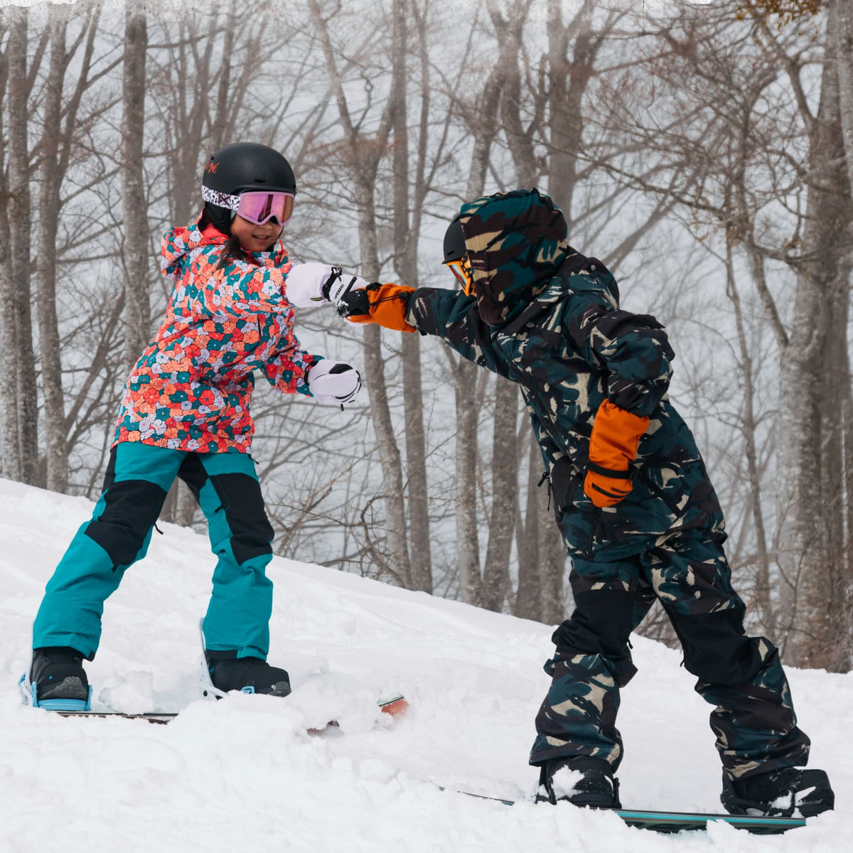 Two children snowboarding on a snowy slope with bare trees in the background. One child wears a colorful floral snowsuit and the other wears a dark camouflage snowsuit with bright orange gloves.