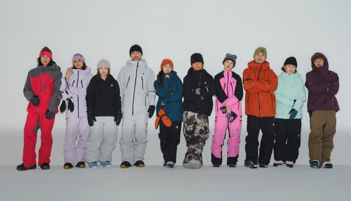 A group of ten people standing in a row wearing various colorful ski suits and jackets against a plain white studio background.