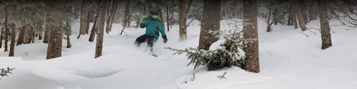 A person snowboarding through deep powder snow in a wooded forest setting, wearing a bright teal jacket, black pants, gloves, and goggles.