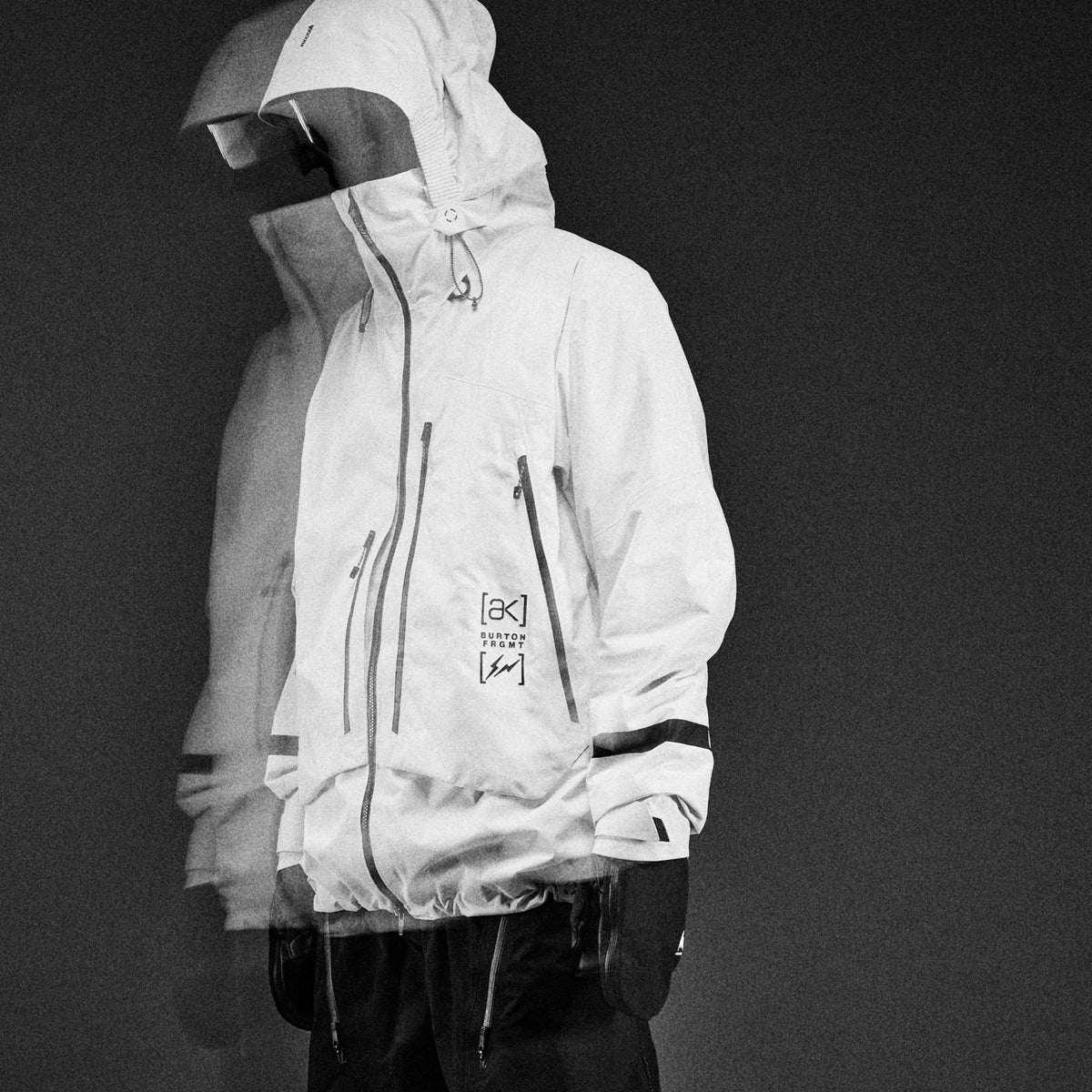 A person wearing a snowboard jacket with multiple zippered pockets and a small printed logo on the chest, shown against a dark studio background.