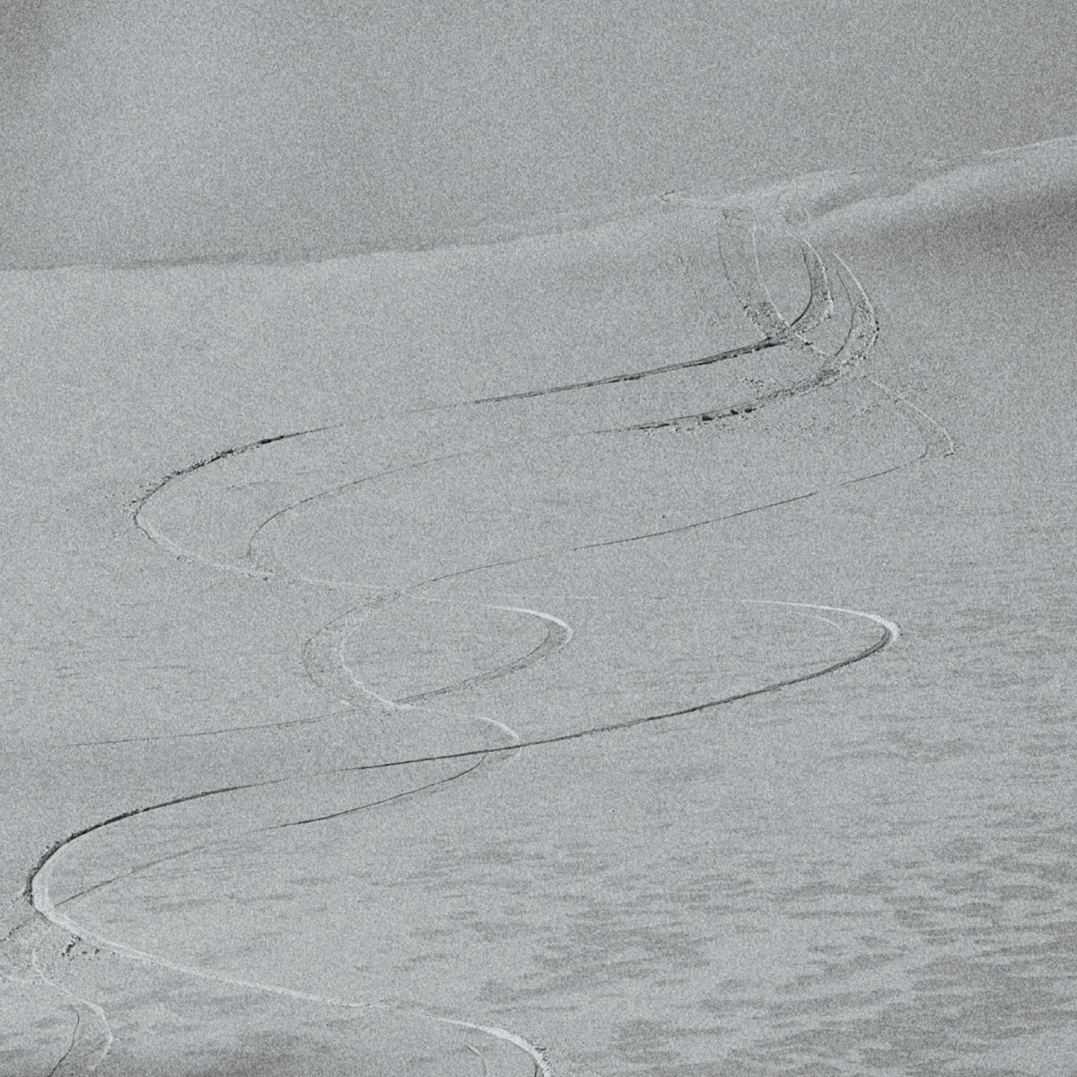 A square photographic print depicting a monochrome gray snowy landscape with winding tire tracks across the snow.