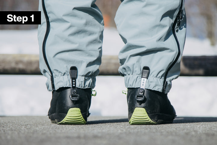 Close-up outdoor shot of a person's lower legs wearing light gray snow pants and black boots. The pants are tucked into a clip on the back of the boots. A black 'Step 1' label appears in the top-left corner.