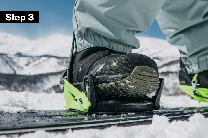 Close-up of a snowboarder's foot in a black snowboard binding with bright neon green accents, standing on a snowboard in the snow with mountains and blue sky in the background. A black label in the top-left corner reads 'Step 3'.