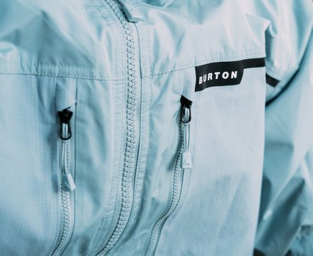 Close-up of a light blue Burton jacket with a black Burton logo patch on the chest and two zippered pockets.
