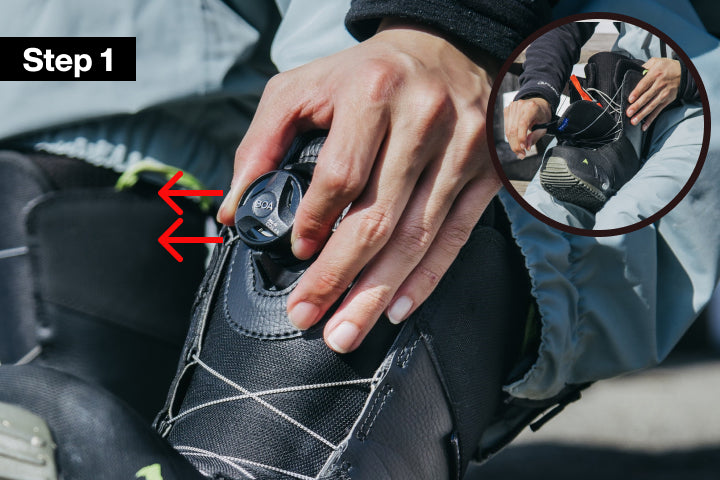 Instruction graphic showing how to fasten a black snowboard boot, labeled 'Step 1' with red arrows pointing to the buckle area. A hand is shown adjusting the boot, and a circular inset shows a wider view of the boot and hands.
