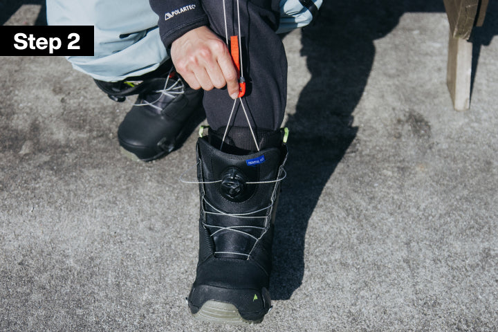 A close-up instructional photo showing a person pulling tight the inner laces while pushing down on the red lace tab of a snowboard boot 