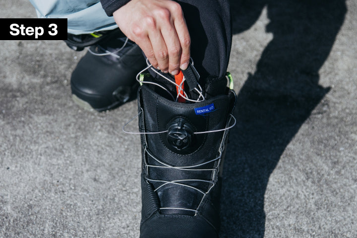 A close-up of a black BOA snowboard boot with a person's hand tucking the inner laces between the outer boot and inner boot liner. Set outdoors on a gray asphalt surface. A black overlay in the top-left corner reads 'Step 3'.