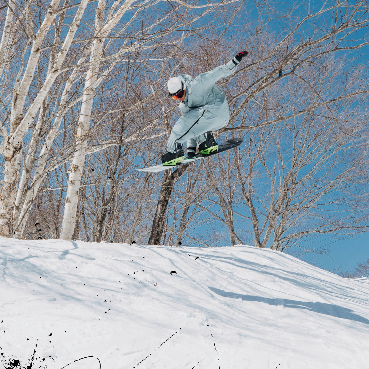 A snowboarder in a light gray outfit jumps through the air on a snowy slope, with bare birch trees and a clear blue sky in the background.