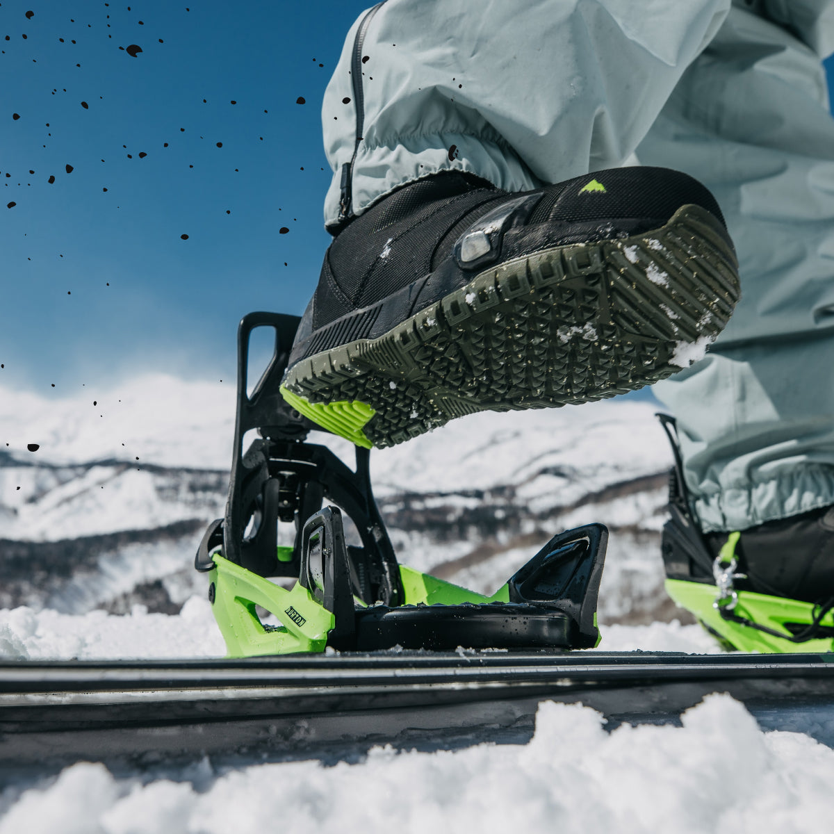 Close-up action shot of a snowboarder's foot in a black snowboard binding with bright neon green accents, set against a snowy mountain background under a clear blue sky.