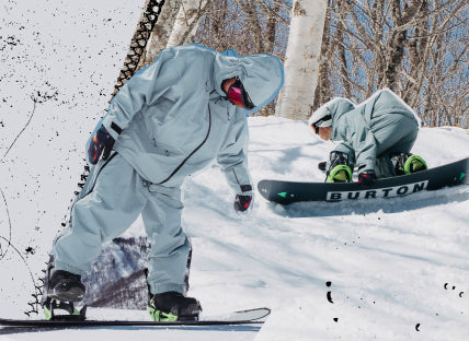 Two snowboarders in light gray snow suits on a snowy mountain slope, with one stepping on to their board and one and one riding.