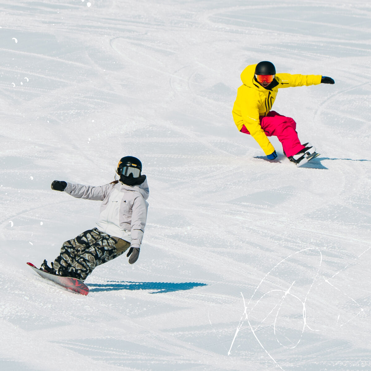 Two snowboarders on a snowy slope. One snowboarder in the foreground wears a light gray jacket, camouflage pants, and a black helmet.