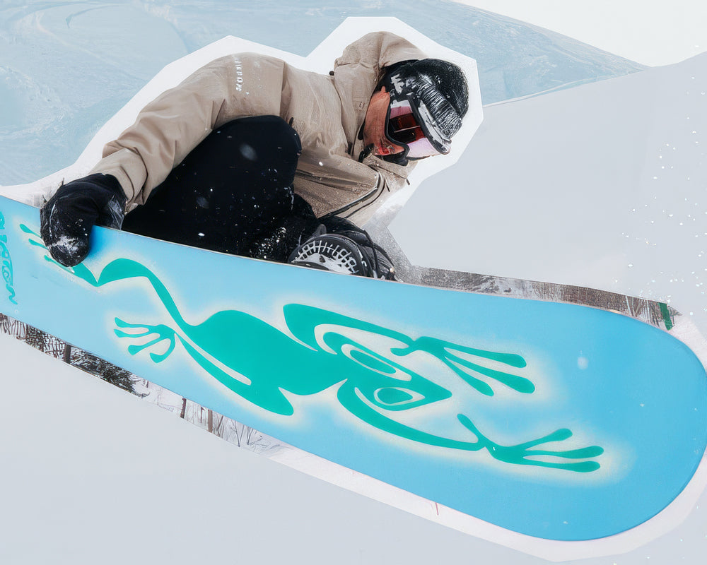 snowboarder in a beige jacket and helmet jumps in mid-air with a light blue Burton snowboard featuring a teal graphic.