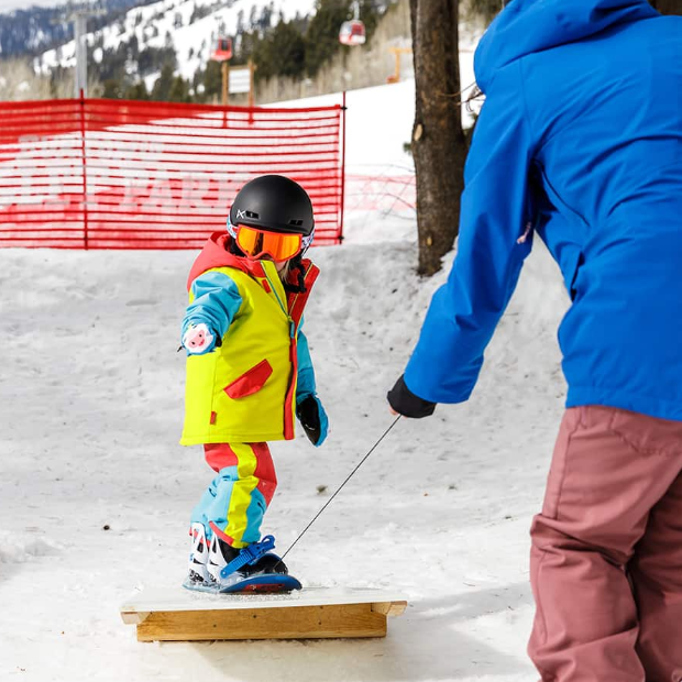 Child in colorful snow gear being taught to snowboard on a Riglet snowboard