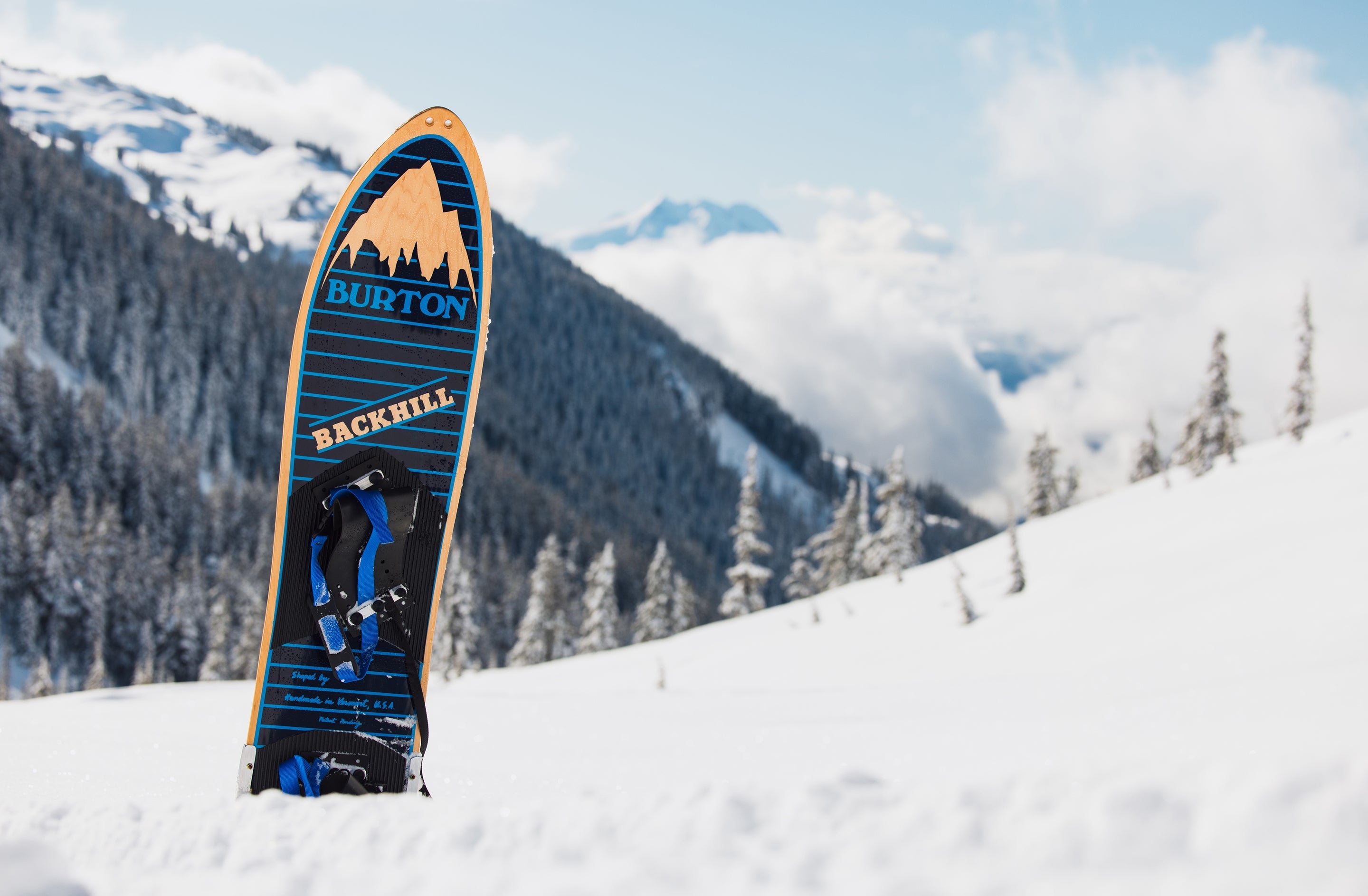 A Burton Backhill snowboard standing upright in a snowy mountain landscape under a blue sky with clouds.
